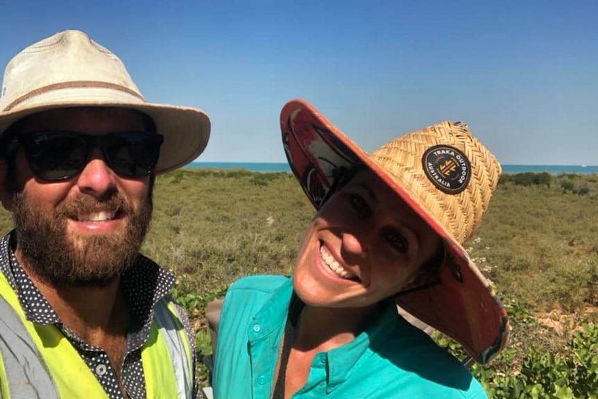 A selfie of a man and woman in hats in front of mangroves and the sea. 
