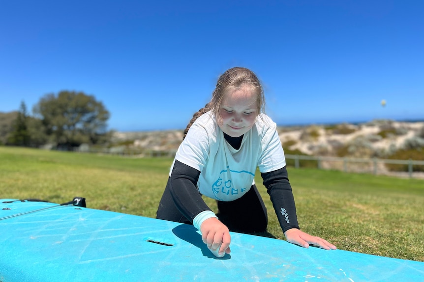 Ruby Taylor on the ground waxing her blue surfboard on a sunny summer's day.
