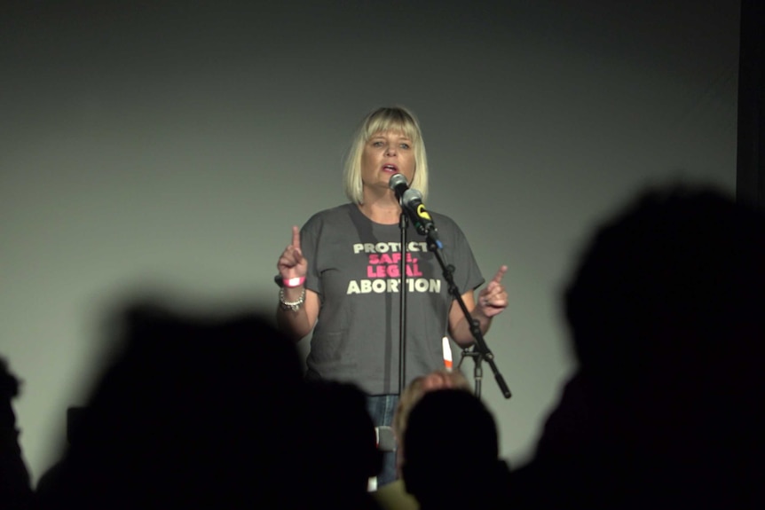 A woman standing on a stage wearing a "Protect safe legal abortion" t-shirt
