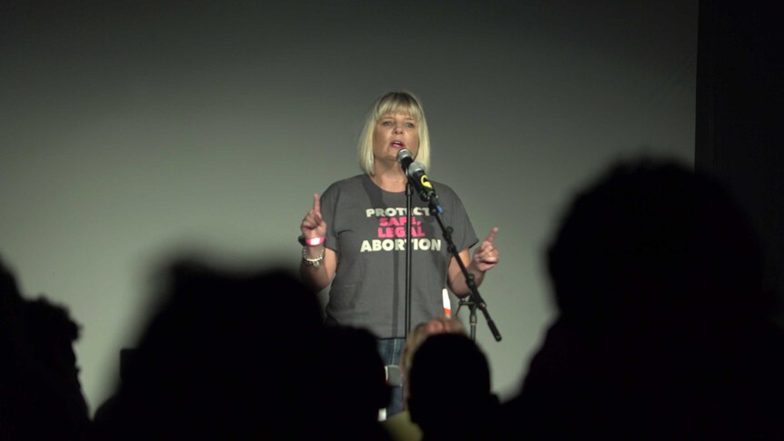 A woman standing on a stage wearing a "Protect safe legal abortion" t-shirt