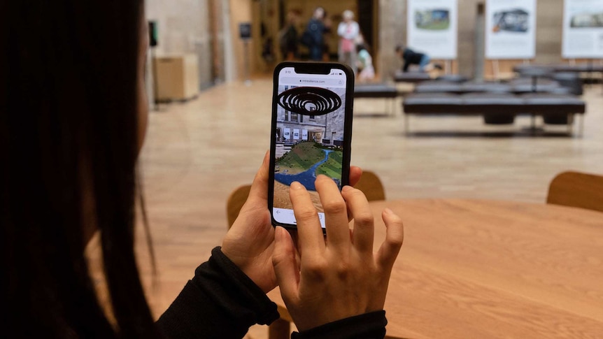 Looking over the shoulder of a woman holding up her phone in a museum. An augmented reality, mountaintop town is on her screen.