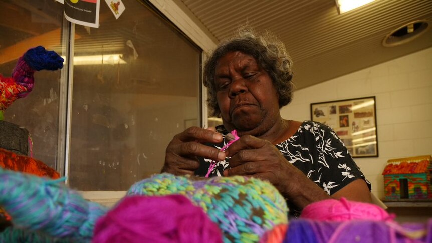 An Aboriginal woman sits at a table knitting. In front of her are brightly coloured balls of wool