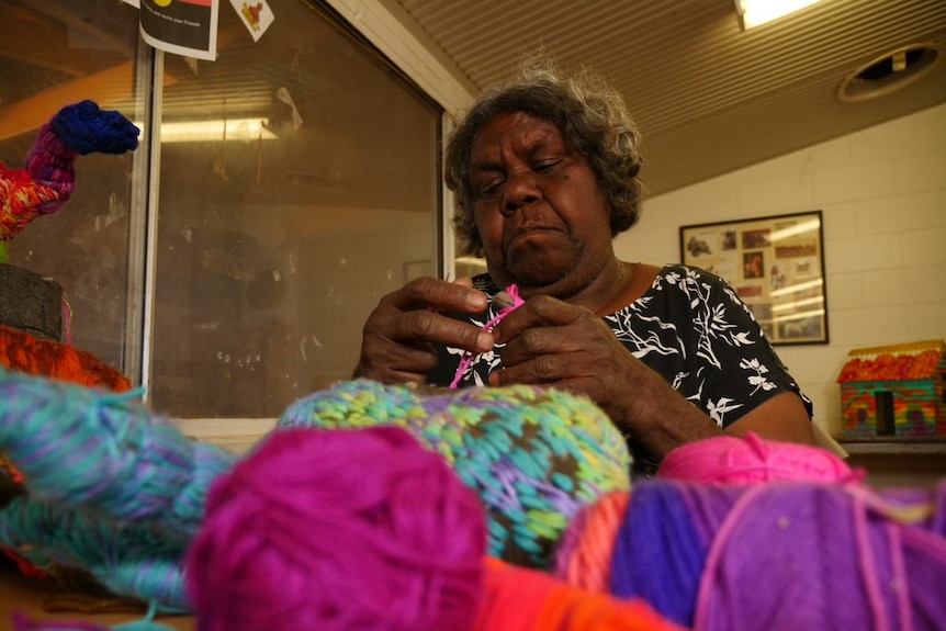 An Aboriginal woman sits at a table knitting. In front of her are brightly coloured balls of wool