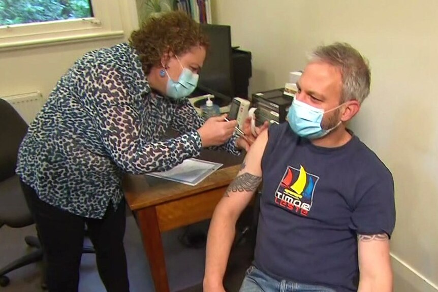 A woman gives a masked man in a t-shirt and jeans a needle.