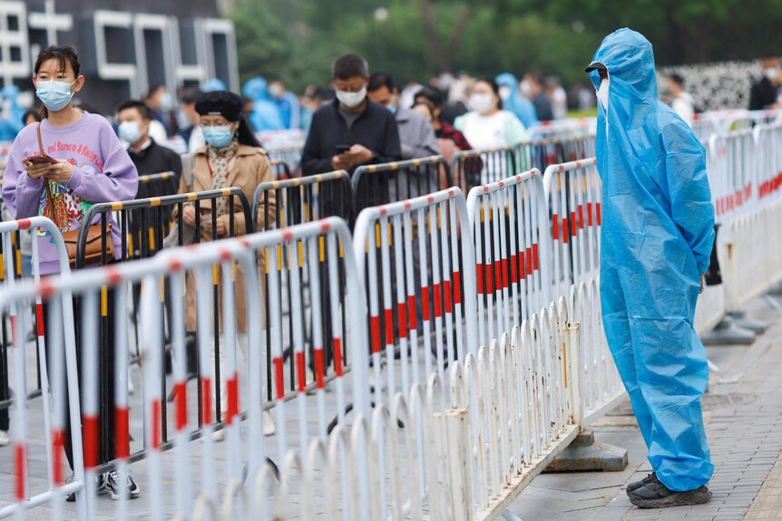 Мужчина в полном костюме СИЗ стоит возле очереди людей за барьером. 