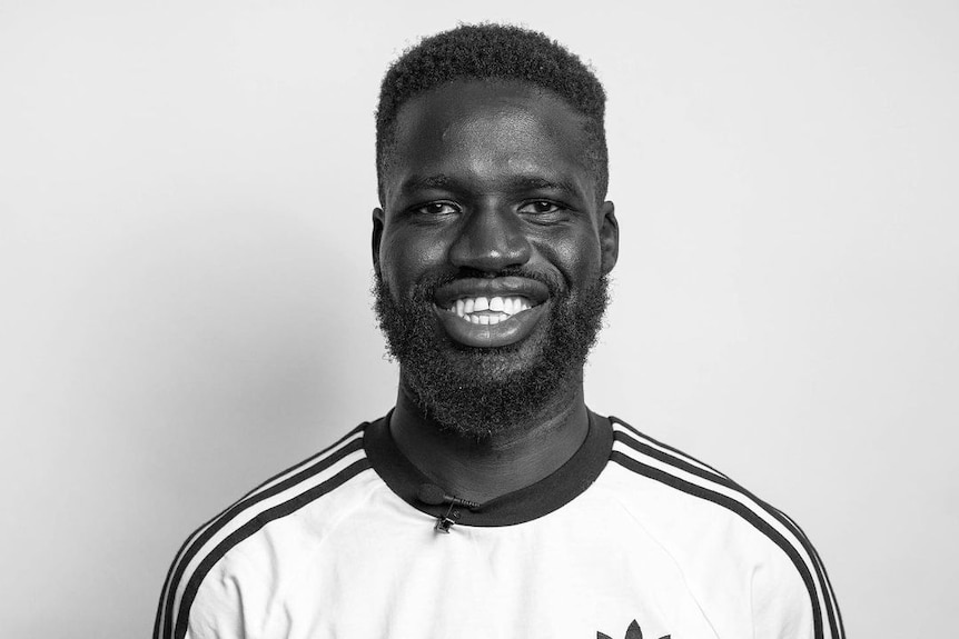 A man with dark short hair wearing a white T-shirt smiling in a black and white photo