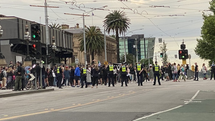 High-visibility-vested police officers stand in the middle of the street as dozens of protesters march across the road.