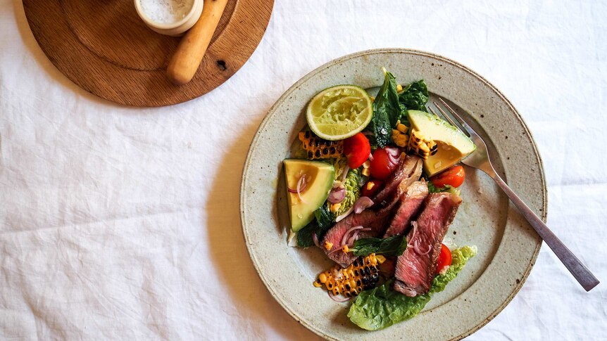 A plate of steak salad with medium rare sliced steak, cos lettuce, charred corn, lime and avocado, a fast warm weather dinner.
