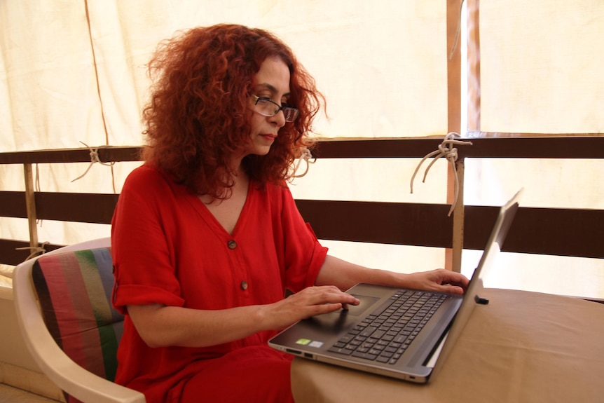 A woman with red hair sits at a table in a red dress working on a laptop computer.