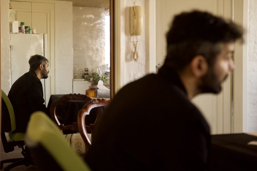 A man sits in his house. His side profile is reflected in a mirror behind him.