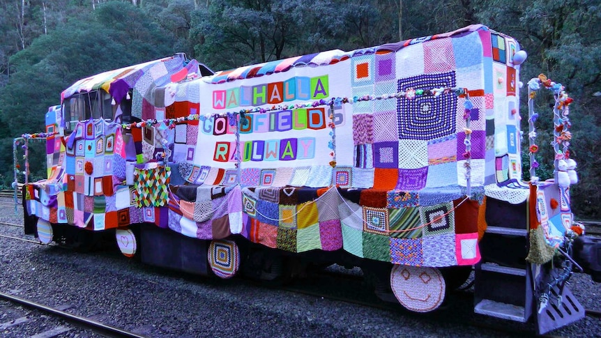 Locomotive engine on rail tracks surrounded by forest, is draped in a  custom-fitted knitted and crocheted patchwork garment.