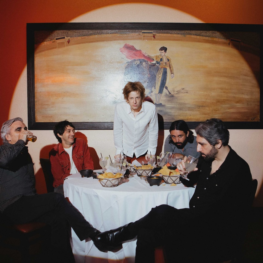 Britt Daniel standing at a table, with 2 members of Spoon seated on each side. Behind is a painting of a matador and a bull.