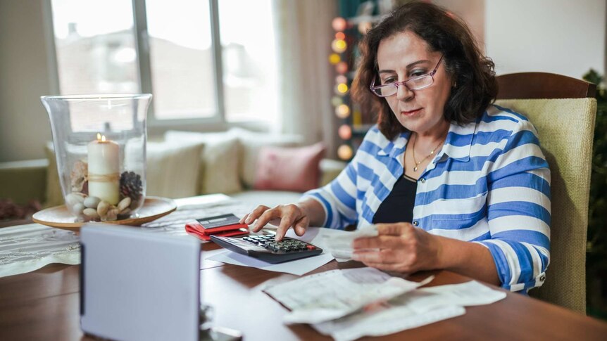 A woman sits at a table with a calculator and bills, running the numbers on her expenses.