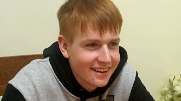 A smiling young man with short ginger hair sits at a table with a birthday cake in front of him