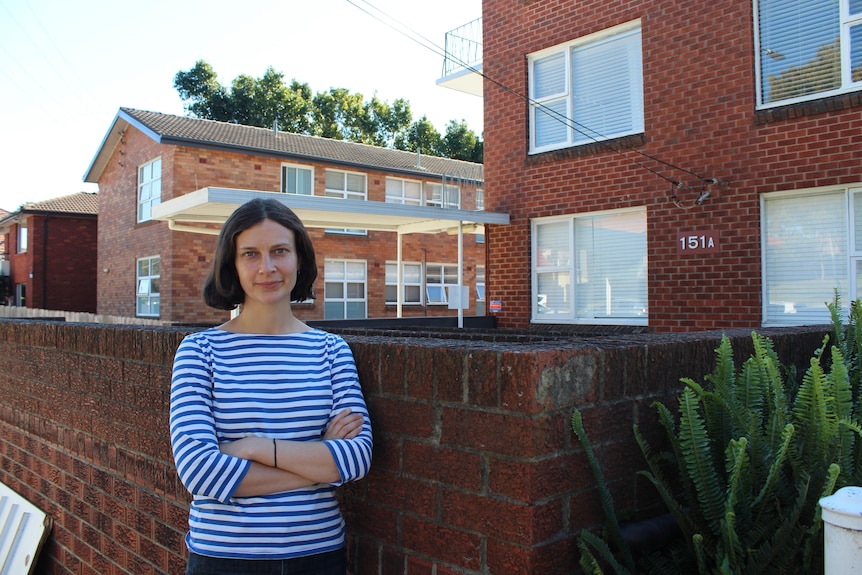 Marlee Bower leans against a brick fence with her arms crossed, infront of a row of brick apartment buildings.