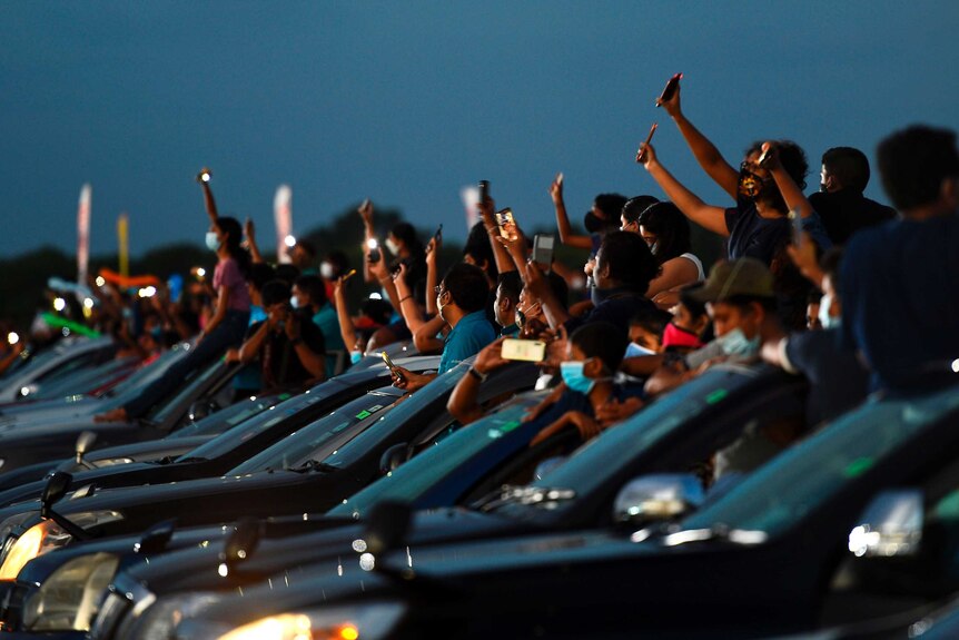 People stand by and hang out of their cars in a row, holding their phones above them to film a live concert.