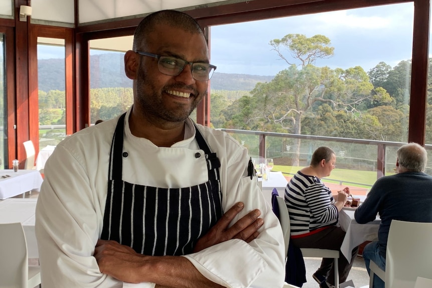 Silas Masih stands smiling in the restaurant, with large glass windows and trees in the background.