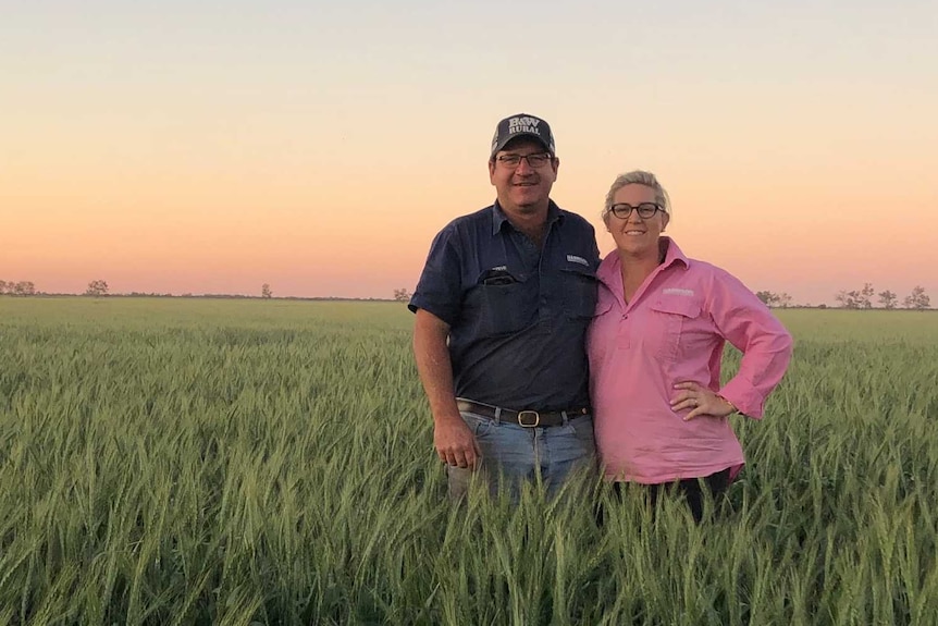 A man and woman stand in a field with their arms around one another.