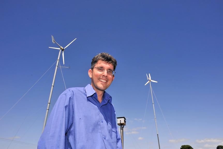 A man smiles. He is standing in front of two wind turbines.