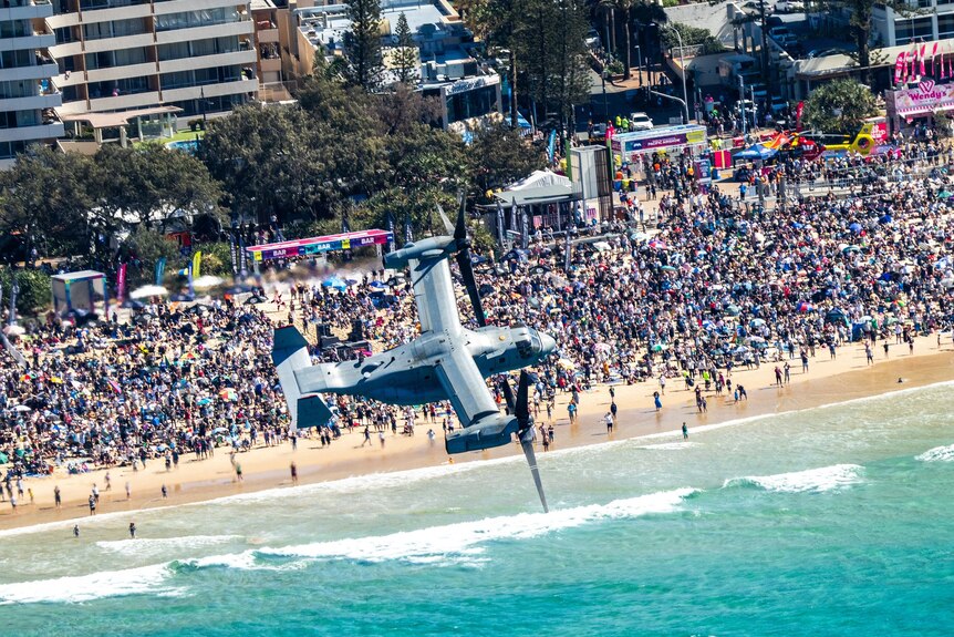 a military aircraft flies low over a beach filled with spectators