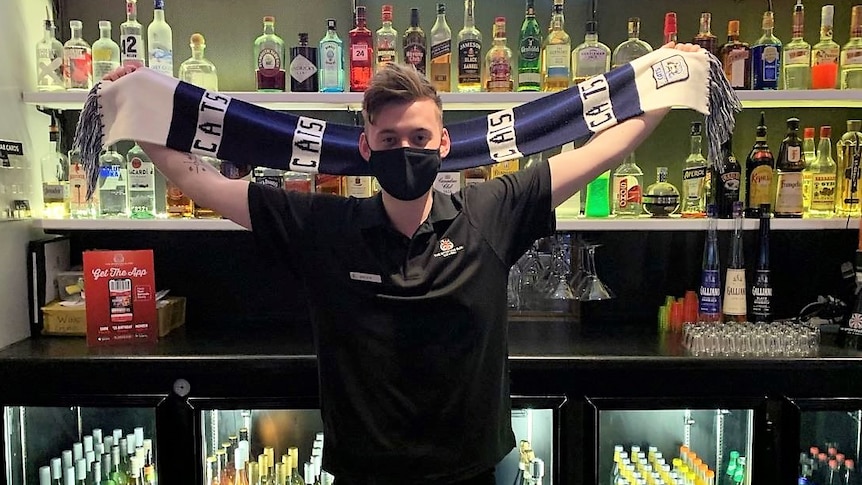 A young man stands behind a bar holding a blue and white football scarf. Alcohol bottles line the shelf behind him.