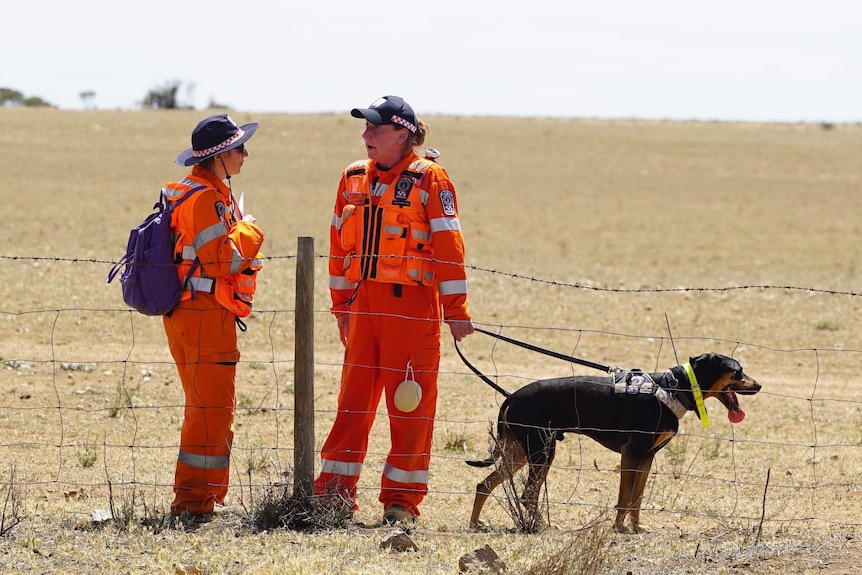 Two women in orange coveralls with a dog on a lead in front of a broadacre farm