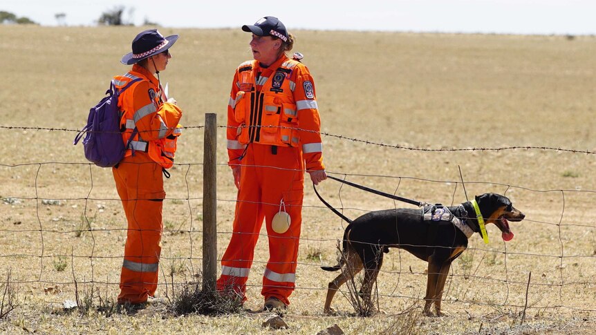 Two women in orange coveralls with a dog on a lead in front of a broadacre farm