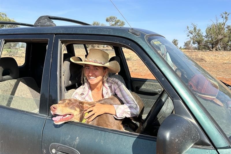 A woman in a cowboy hat sits in the front seat of her car with a brown kelpie.