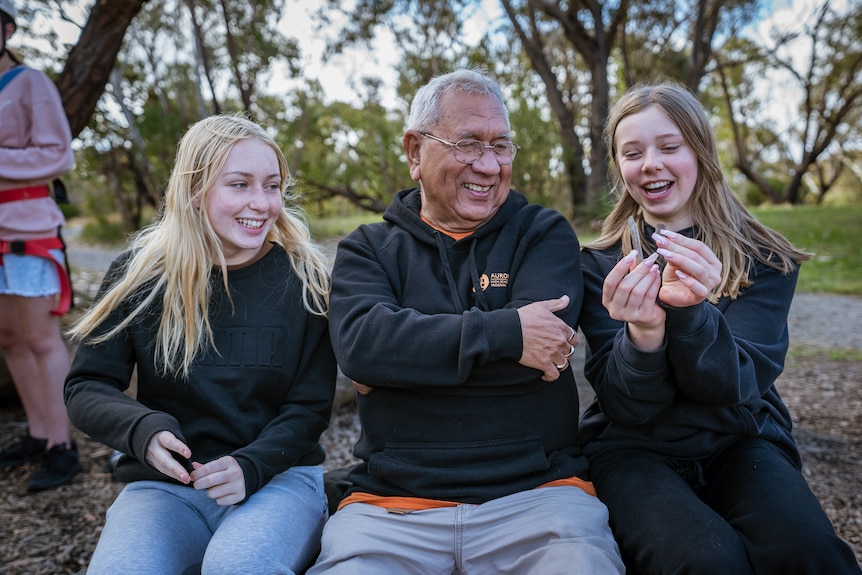 Two young girls sit next to a man with grass and trees behind them. One of the girls shows the man something and they smile.