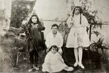 black and white image of four children in fancy dress, all look sad. three are standing and one sits in the middle.