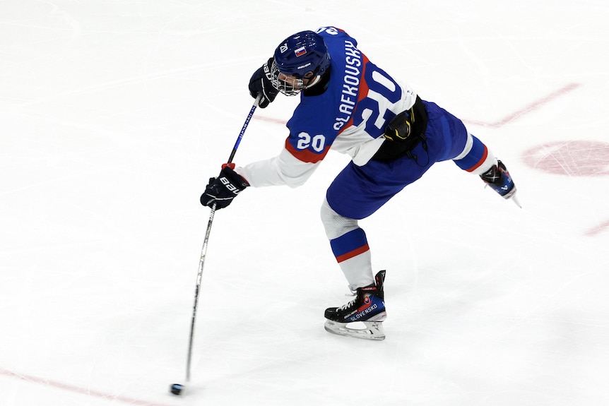 A Slovakian ice hockey player balances on one skate as he bends his stick at the second of impact for a scoring shot.