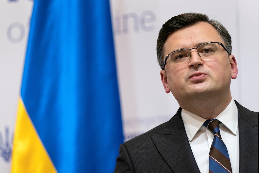A tilted mid-shot of dark-suited man wearing rimless glasses who is standing in front of a Ukrainian flag.
