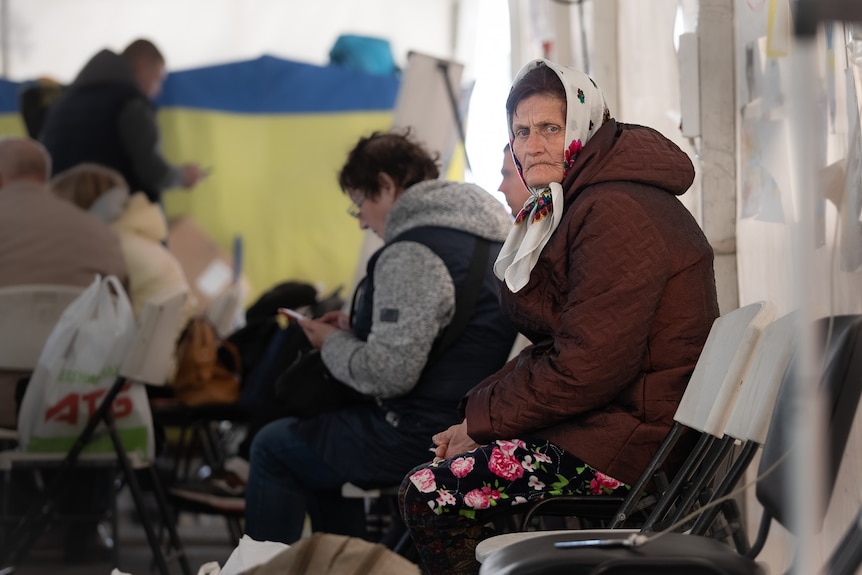 A woman wearing a hijab, large coat and floral skirt sits on a chair with other people gathered in a room.