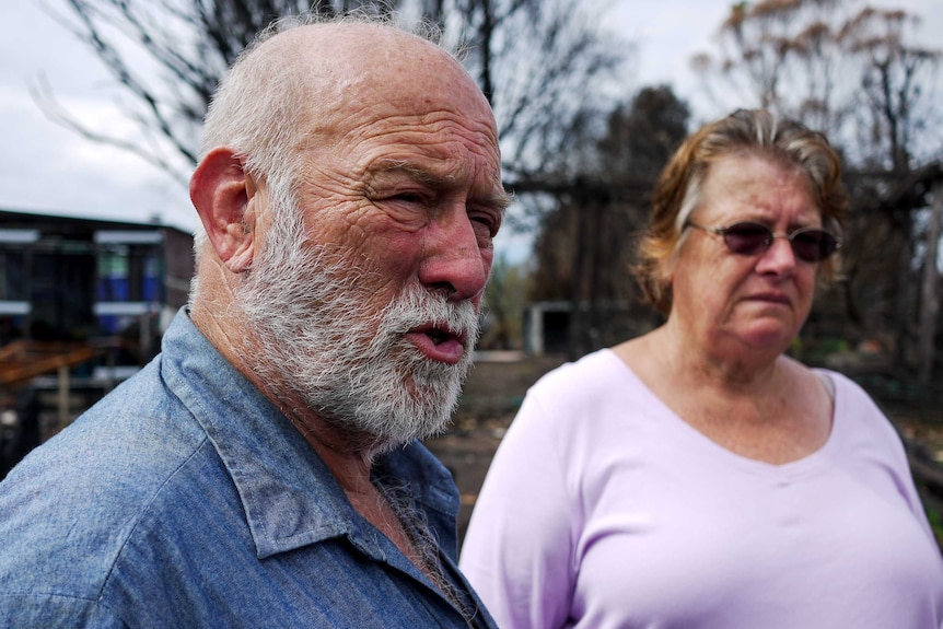 Man with grey beard and blue shirt and woman in purple t-shirt wearing glasses stand outside with concerned look on their faces