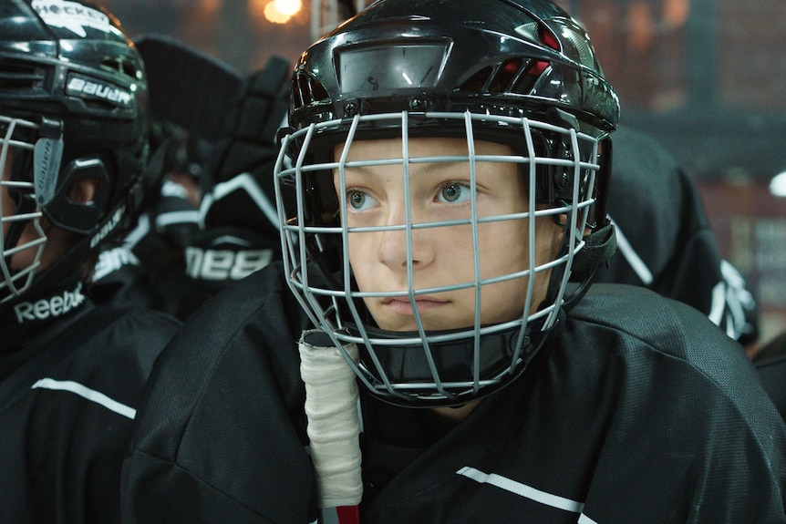 A teenage boy sits looking determined. He is wearing a black helmet and a hockey uniform. 