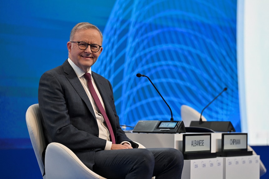 Anthony Albanese smiles as he sits on a stage with a blue background