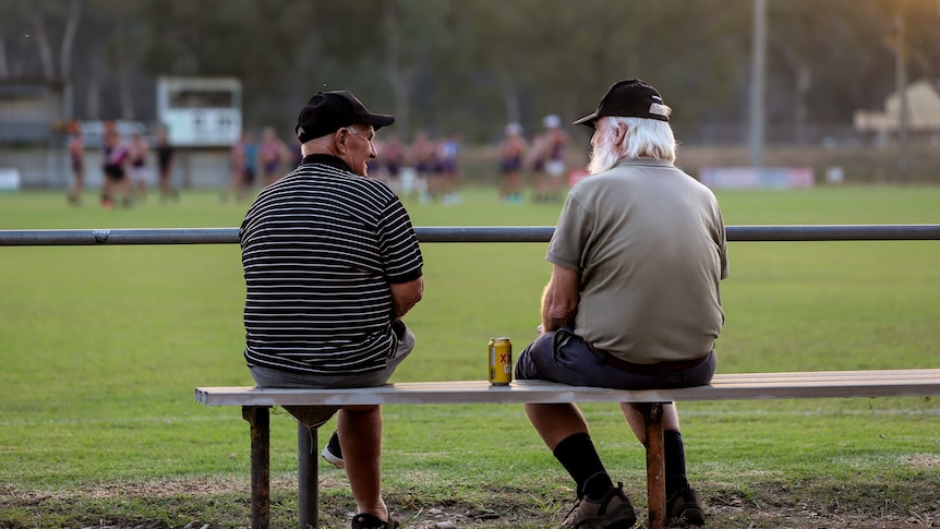 Two older men in conversation sitting on a bench on the boundary of a football oval, with players in distance under sunset
