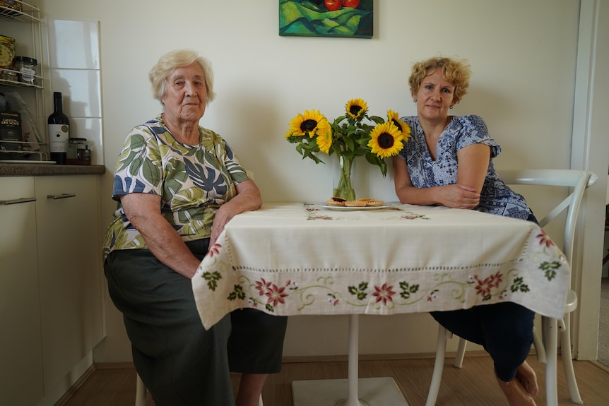 Elderly woman and her daughter are sitting at a dining table with a vase of sunflowers.