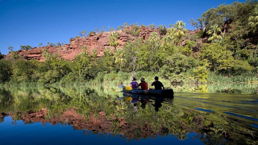 Three people paddle a canoe across a glassy waterhole surface reflecting blue sky, bare rock and lush bush onthe shore's edge.