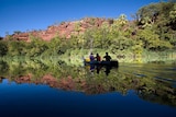 Three people paddle a canoe across a glassy waterhole surface reflecting blue sky, bare rock and lush bush onthe shore's edge.
