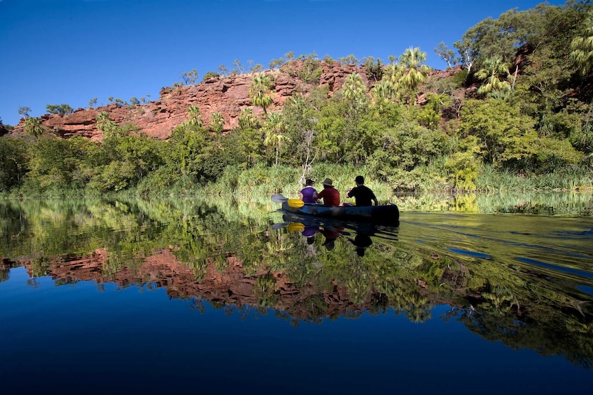 Three people paddle a canoe across a glassy waterhole surface reflecting blue sky, bare rock and lush bush onthe shore's edge.