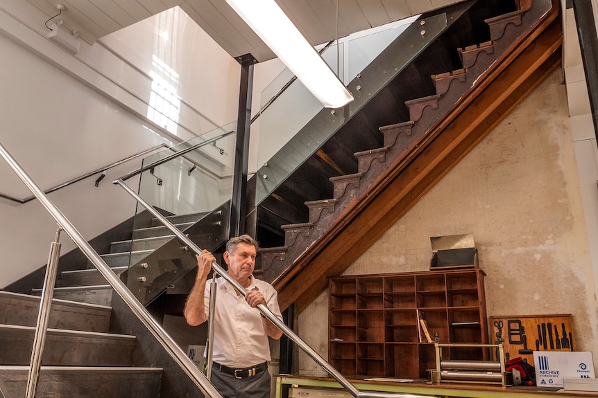 A man stands below an ornate timber staircase the sits below a modern staircase.