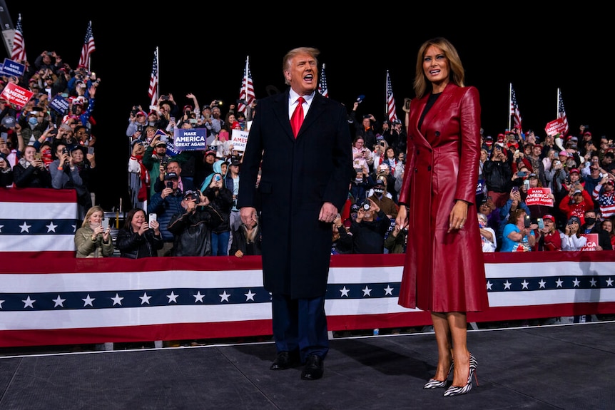 You view Donald Trump and Melania Trump on a rally stage at night.