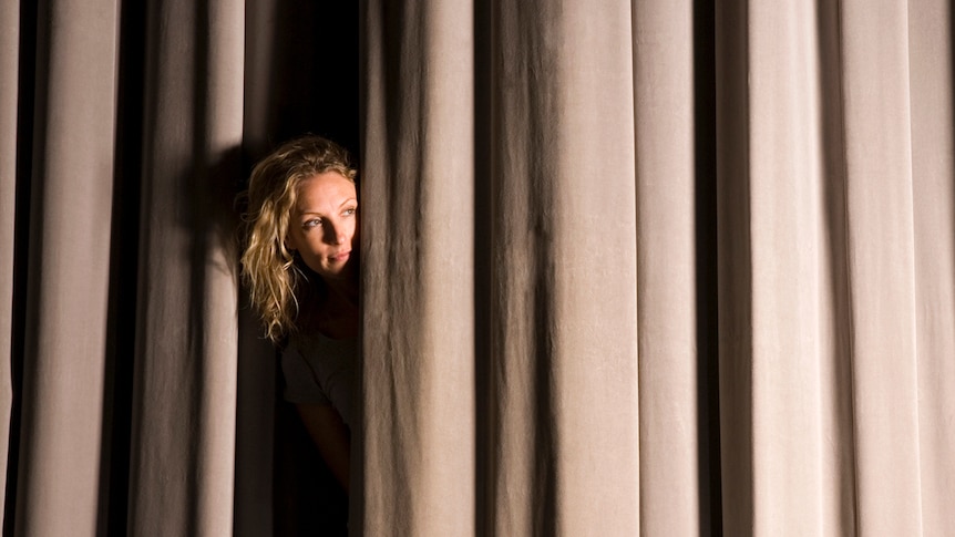 woman with blonde curly hair peeking through light brown velvet stage curtain.