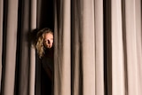woman with blonde curly hair peeking through light brown velvet stage curtain.