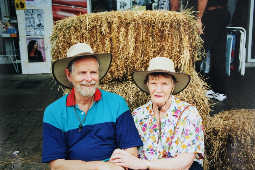 An older couple sit in front of a hay stack smiling with hats on.