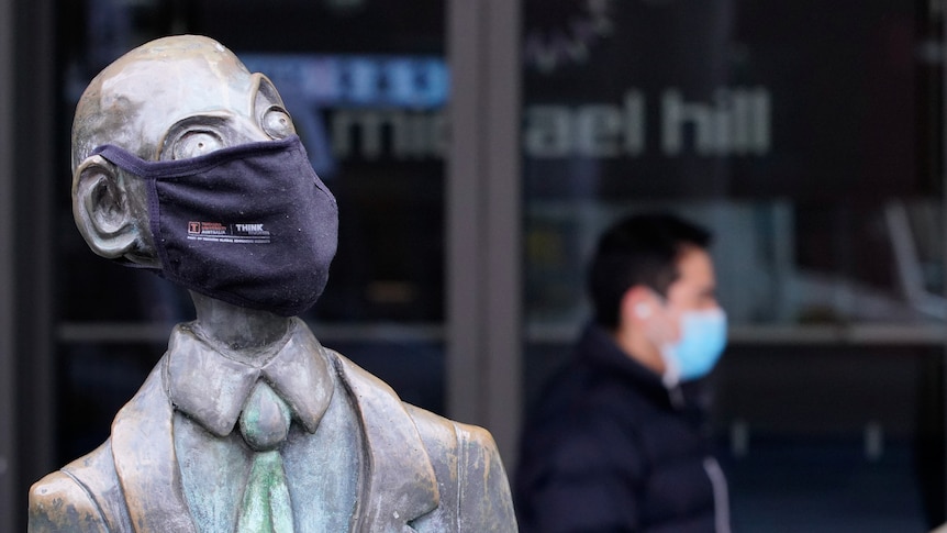 A man wearing a protective face mask walks past a masked statue in Melbourne