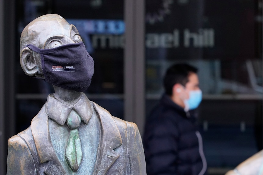 A man wearing a protective face mask walks past a masked statue in Melbourne