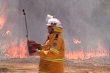 A man in a yellow fire suit and white helmet carries a red pot. Fire burns through scrub in the background.