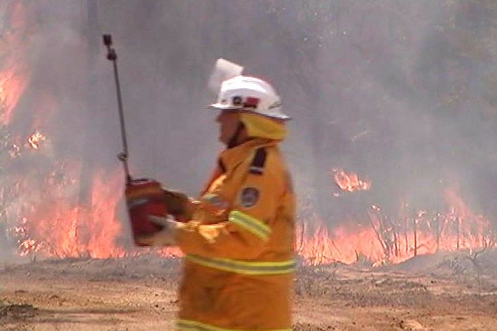 A man in a yellow fire suit and white helmet carries a red pot. Fire burns through scrub in the background.
